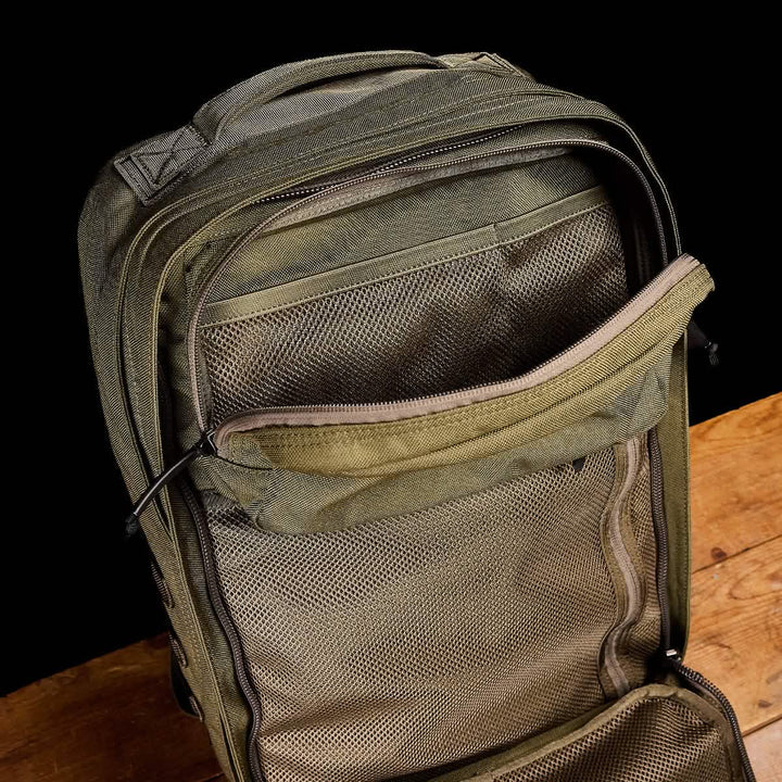 The open GR2 backpack from GORUCK, in green, sits on a wooden surface against a black backdrop. Its Special Forces quality is clear as the unzipped main compartment displays mesh pockets for exceptional organization, making it perfect for those looking for carry-on compliant options.