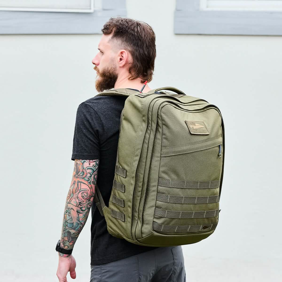 A person sporting a beard and arm tattoos, donned in a black t-shirt, carries the GORUCK GR2 backpack. They stand outside against a pale wall with windows, gazing to the side. The GR2's design guarantees it's both durable and suitable as carry-on for any adventure.