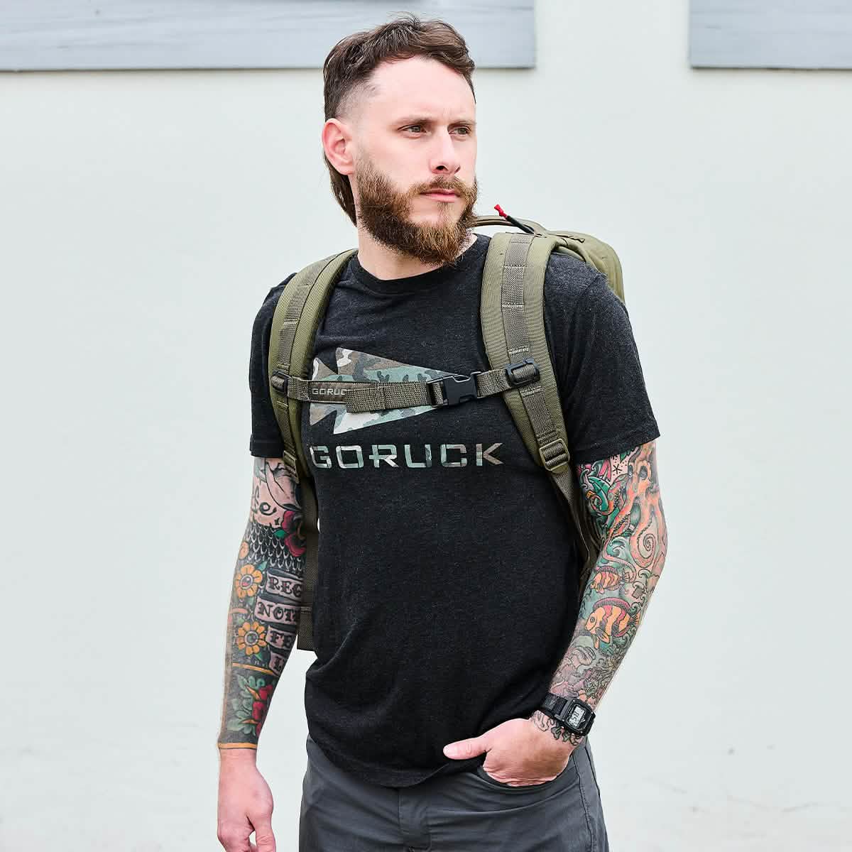 A bearded man with tattoos on both arms stands outdoors, wearing a dark t-shirt featuring a GORUCK graphic. He carries the GORUCK GR2 Special Forces quality green backpack, looking to the left with a neutral expression.