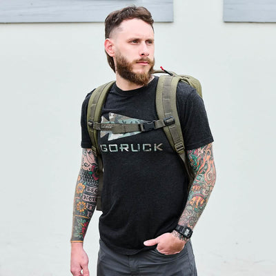 A bearded man with tattoos on both arms stands outdoors, wearing a dark t-shirt featuring a GORUCK graphic. He carries the GORUCK GR2 Special Forces quality green backpack, looking to the left with a neutral expression.