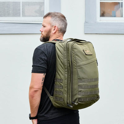 A man with short hair and a beard is wearing a black T-shirt and carrying an olive green GORUCK GR2 - Cordura ruck. He stands sideways, showcasing the backpack's special forces design featuring horizontal strap details. The background is a pale wall with two windows.