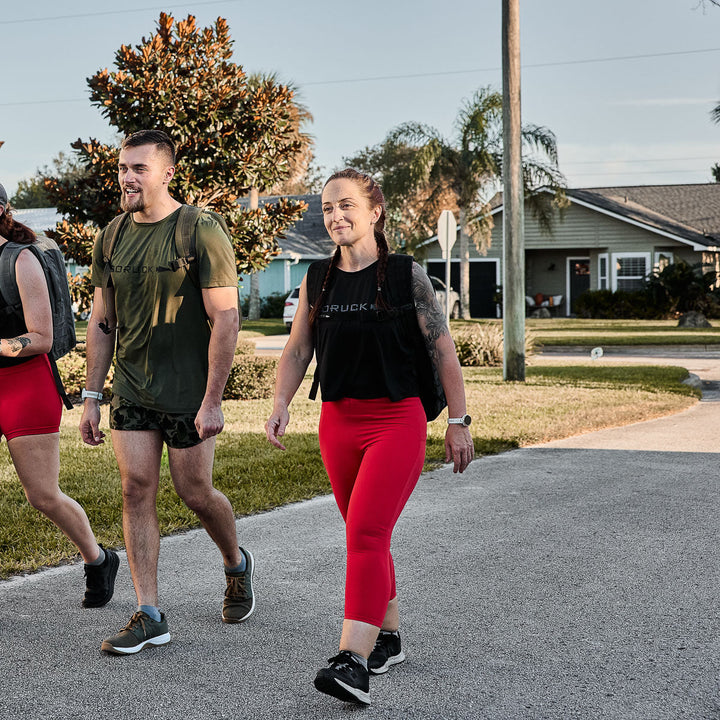 Three individuals walk down a suburban street, backpacks in tow, dressed in athletic gear. The women wear GORUCK Women's Cropped Training Leggings made from ToughFlex fabric, providing squat-proof comfort for their outdoor adventure.