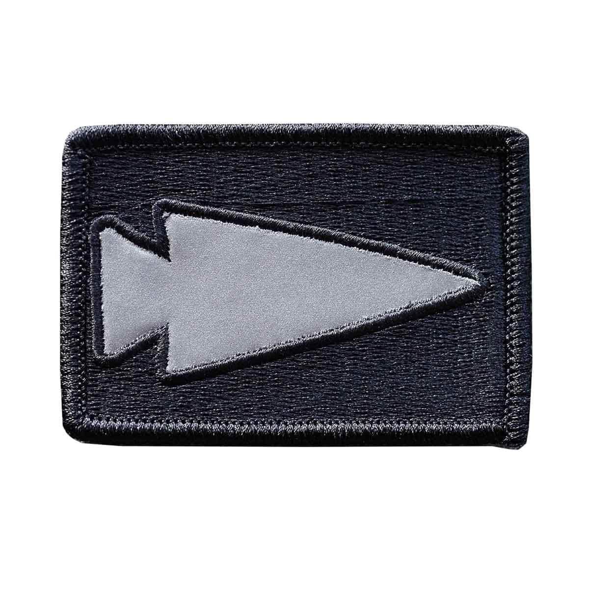 Introducing the GORUCK Patch - Reflective Spearhead: This rectangular black patch showcases an embroidered gray spearhead symbol centered on the patch, oriented to the left. It is designed with a stitched border and equipped with a VELCRO backing for effortless attachment.