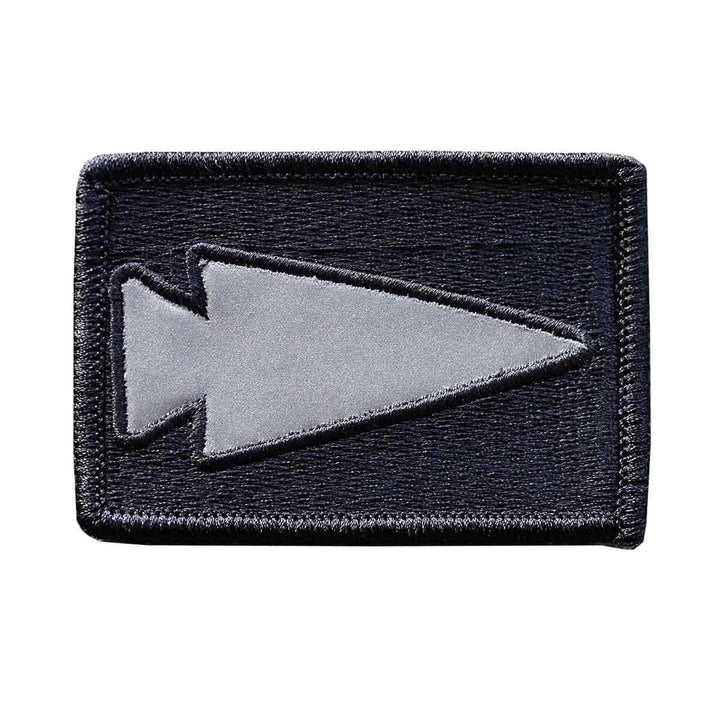 Introducing the GORUCK Patch - Reflective Spearhead: This rectangular black patch showcases an embroidered gray spearhead symbol centered on the patch, oriented to the left. It is designed with a stitched border and equipped with a VELCRO backing for effortless attachment.