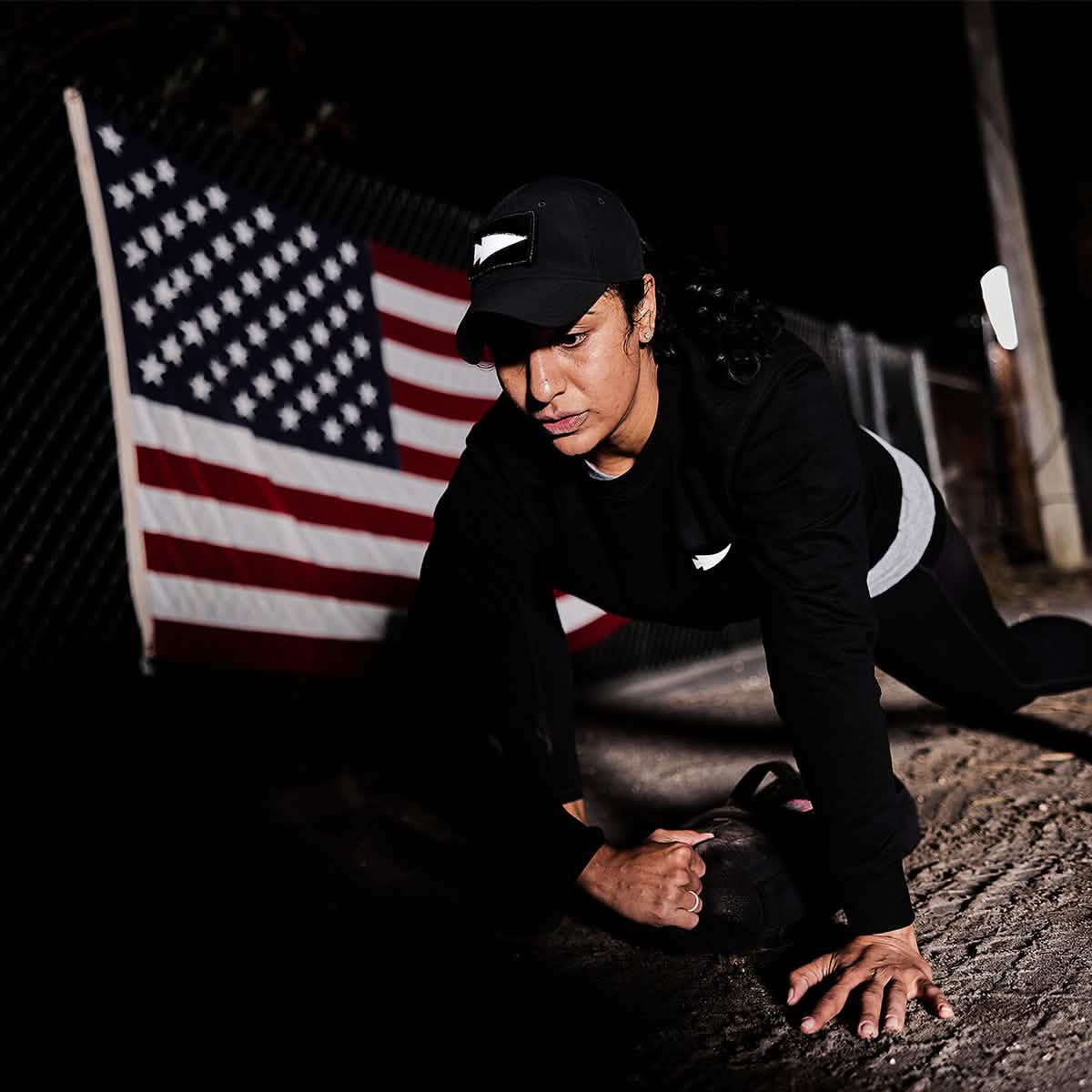 Clad in a GORUCK black cap and sweatshirt adorned with the Reflective Spearhead patch, an individual is exercising with a sandbag outdoors at night. In the backdrop, an American flag is draped over a fence. The dimly lit scene highlights the physical exertion involved.