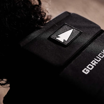 Close-up of a black backpack with an embroidered Reflective Spearhead patch. The GORUCK brand name is visible next to a strip of VELCRO. The background is out of focus, highlighting the backpack's texture and details.