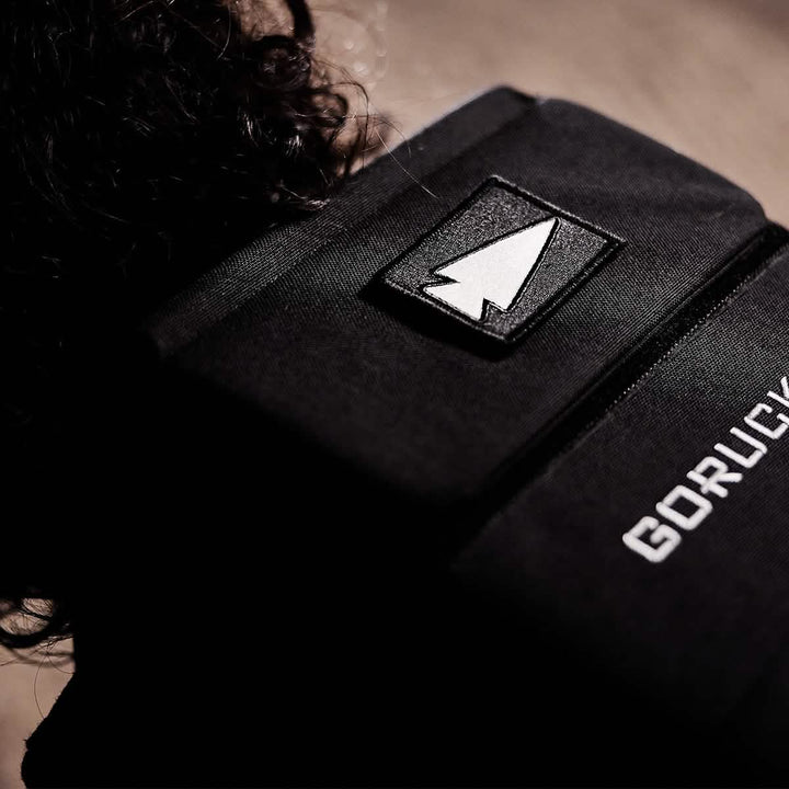 Close-up of a black backpack with an embroidered Reflective Spearhead patch. The GORUCK brand name is visible next to a strip of VELCRO. The background is out of focus, highlighting the backpack's texture and details.