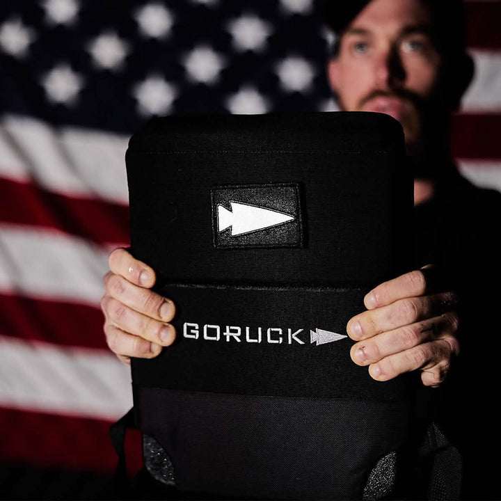 A person holding a GORUCK backpack faces the camera with an American flag in the background. The GORUCK logo and a reflective spearhead patch are visible, adding a touch of rugged flair to the scene.