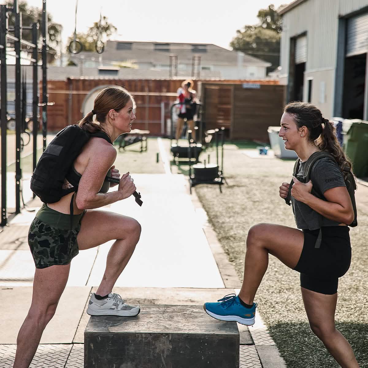 Two women are outdoors, engaging in a rucking workout with backpacks while stepping onto boxes. One sports a gray shirt paired with camouflage shorts, and the other is dressed entirely in black. With a gym setting as their backdrop, their use of GORUCK's Ruck Clubs Starter Kit - Heavy suggests they are preparing for an upcoming GORUCK event.