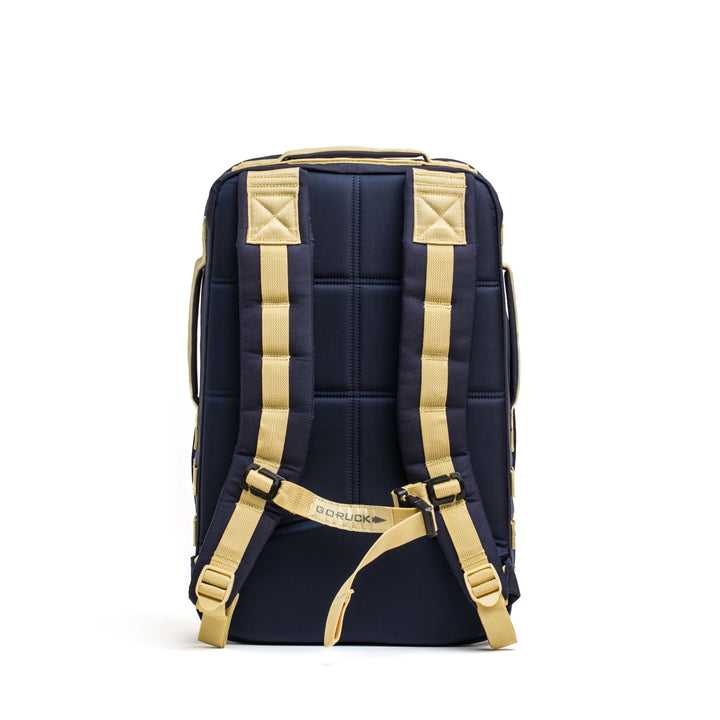 The GORUCK Rucker 4.0 features a navy and beige design with padded straps and a top handle, viewed from the back.
