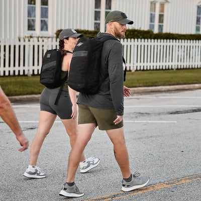 Two individuals sporting GORUCK gear enjoy rucking down a street with their Rucker 4.0 backpacks. The person on the right is dressed in a cap, hoodie, and shorts, while their companion wears a cap, T-shirt, and shorts. A white fence stands behind them.