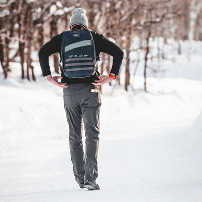 A person wearing the Rucker 4.0, possibly weighted with a Ruck Plate, walks on a snowy path flanked by trees—a classic winter rucking scene viewed from behind.
