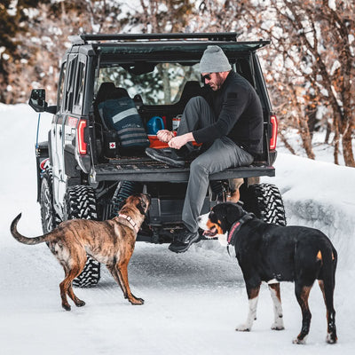 A man sits on the tailgate of an SUV in the snow, pulling on boots with two dogs at his side—ready for a rucking adventure with his reliable Rucker 4.0.
