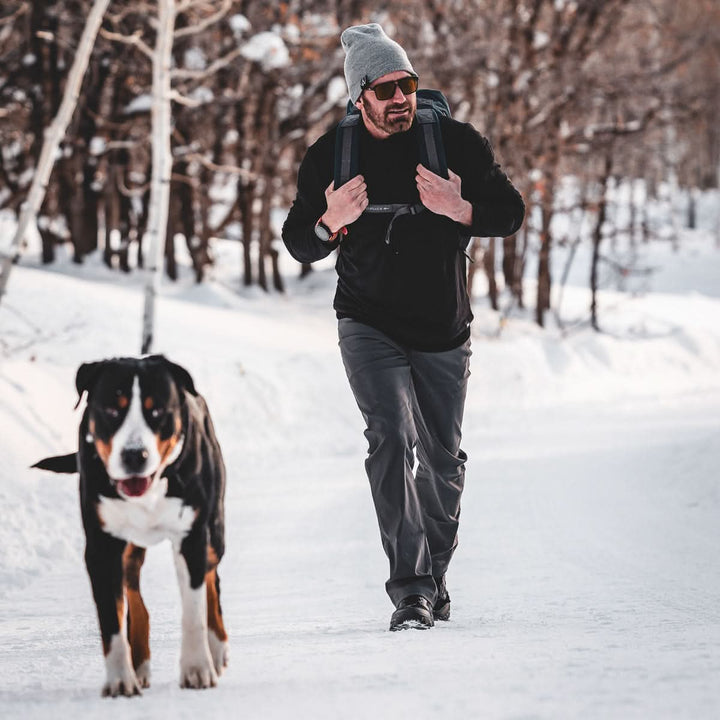 A man carrying the Rucker 4.0 backpack walks on a snowy path with a large dog, surrounded by trees.