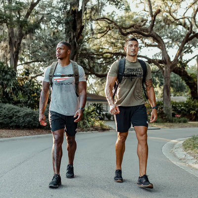 Two men stroll along a tree-lined road, dressed in athletic gear and carrying backpacks loaded with the GORUCK Ruck Clubs Starter Kit - Light. They appear focused and fit, relishing an invigorating rucking session under the shade of large trees.