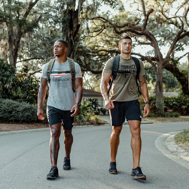 Two men stroll along a tree-lined road, dressed in athletic gear and carrying backpacks loaded with the GORUCK Ruck Clubs Starter Kit - Light. They appear focused and fit, relishing an invigorating rucking session under the shade of large trees.