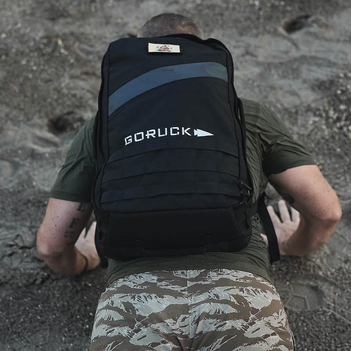 A participant in a GORUCK event is seen crawling on sandy ground while wearing the Ruck Clubs Starter Kit - Light by GORUCK. They are dressed in a green shirt and camouflage pants, an ideal outfit for rucking adventures. The backpack prominently displays the "GORUCK" logo, symbolizing the challenge and endurance demanded outdoors.
