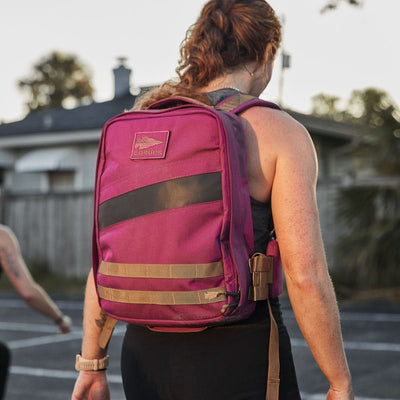 A woman stands outdoors, facing away, wearing a sleeveless top and carrying the Rucker 4.0, a maroon backpack with reinforced construction.