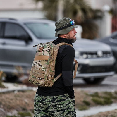 A man in outdoor gear stands near parked cars on a street, ready for a day of rucking with his camouflage Rucker 4.0 backpack and hat.