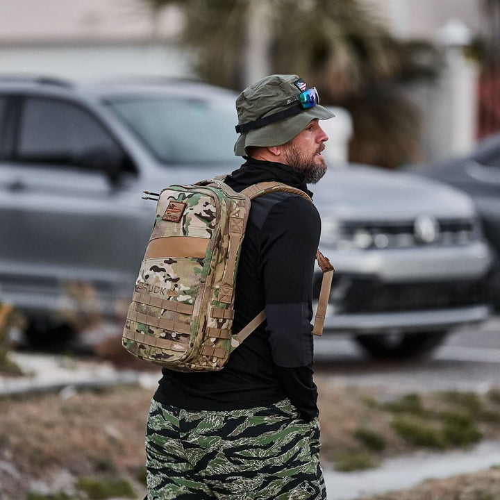 A man in outdoor gear stands near parked cars on a street, ready for a day of rucking with his camouflage Rucker 4.0 backpack and hat.