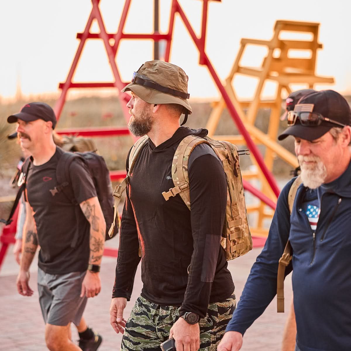 Three men in casual athletic wear walk outside near large red and yellow lifeguard chairs, each carrying their Rucker 4.0 packs, ready for rucking.