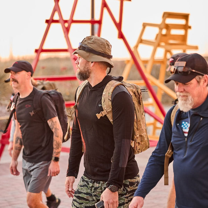 Three men in casual athletic wear walk outside near large red and yellow lifeguard chairs, each carrying their Rucker 4.0 packs, ready for rucking.
