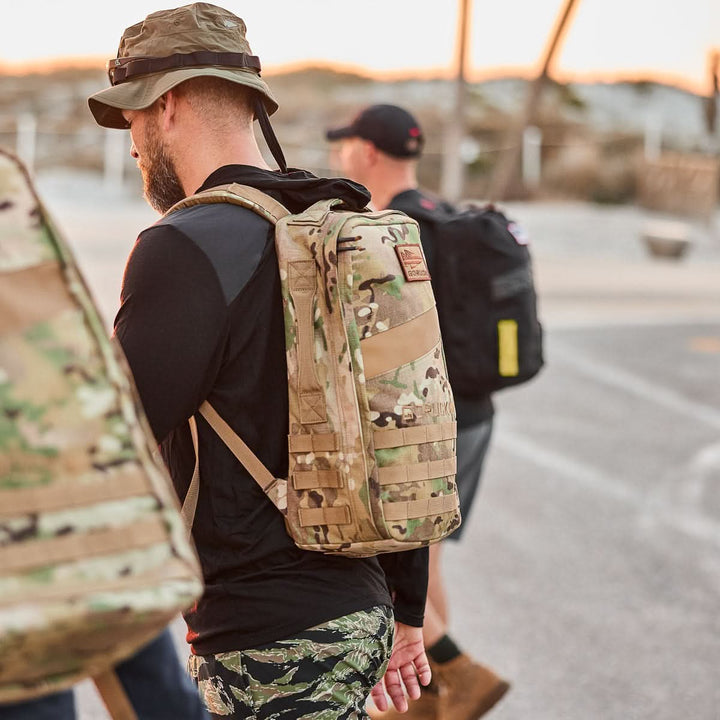 A man wearing a camouflage Rucker 4.0 backpack and hat walks outdoors with others in casual athletic gear, ready for a day of rucking.