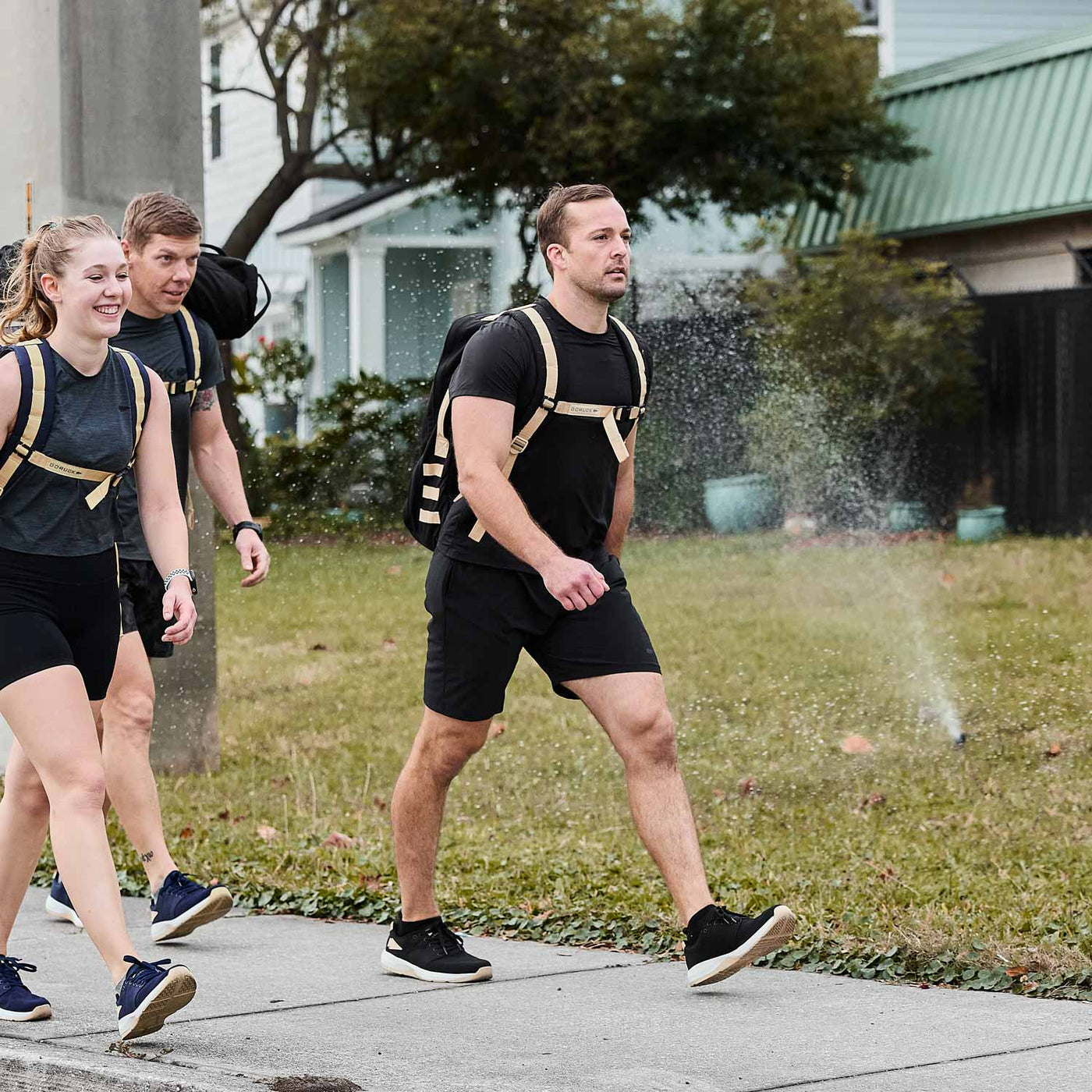 Three individuals carrying GORUCK Rucker 4.0 backpacks are rucking on a sidewalk, passing by a sprinkler that is watering the lawn.