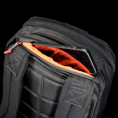 The GR1 Shooter 26L - Built by Scars backpack is made from rugged CORDURA®, features an orange lining, and has a zippered pocket near the shoulder straps for convenient phone storage.