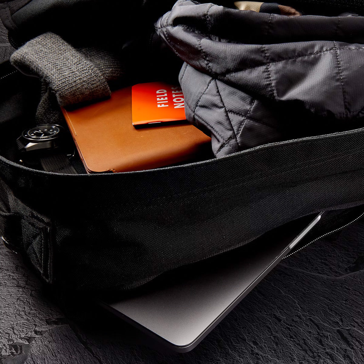 Open black GR1 USA Slick - Ballistic Nylon Cordura backpack with a jacket, notebook, watch, and laptop on a dark textured surface—built with Special Forces-grade quality and a bombproof laptop compartment.