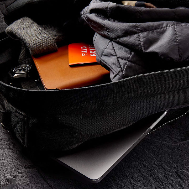 Open black GR1 USA Slick - Ballistic Nylon Cordura backpack with a jacket, notebook, watch, and laptop on a dark textured surface—built with Special Forces-grade quality and a bombproof laptop compartment.