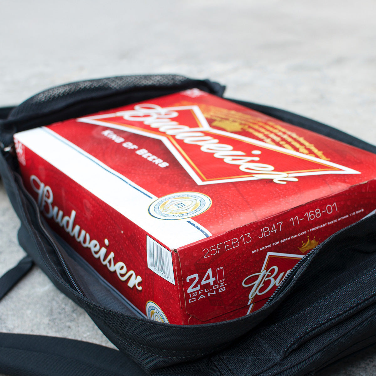 A 24-pack of Budweiser beer in a red box sits inside an open black GR1 USA Slick - Ballistic Nylon Cordura backpack on a concrete surface.