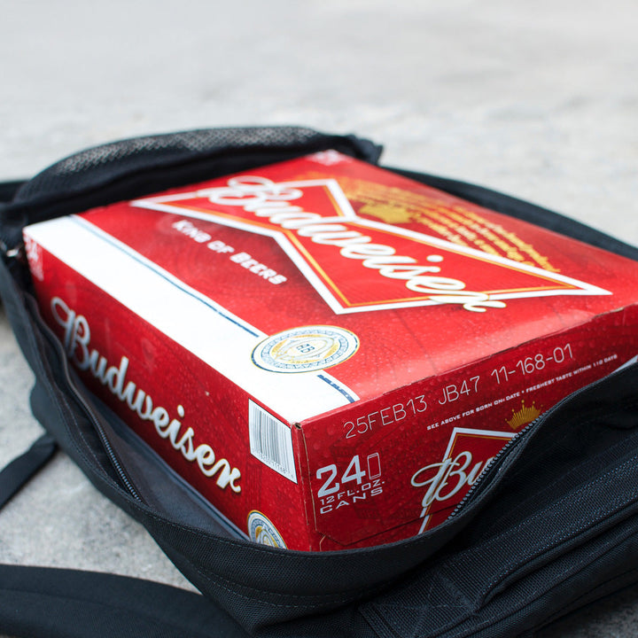 A 24-pack of Budweiser beer in a red box sits inside an open black GR1 USA Slick - Ballistic Nylon Cordura backpack on a concrete surface.