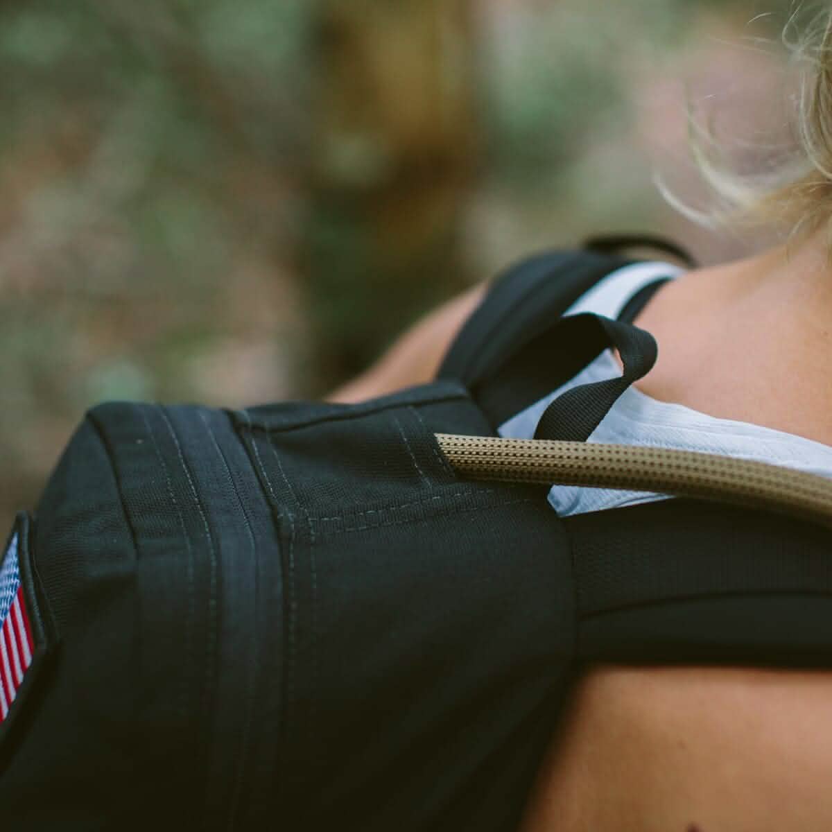 A close-up of someone carrying a black backpack adorned with an American flag patch highlights the "Hydration Bladder" by Source. A tube from this advanced hydration system gracefully drapes over their shoulder. The design, taking cues from Special Forces gear, incorporates antimicrobial technology to ensure enhanced freshness against a backdrop of blurred greenery.