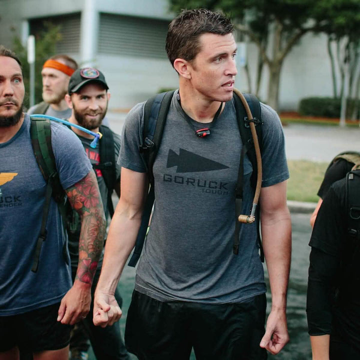 A group of men in athletic gear stands together outdoors, poised for a physical challenge. The man at the center wears a GORUCK Tough shirt and carries a backpack featuring a Source Hydration Bladder, embodying the readiness of Special Forces. Greenery and a building form the backdrop to their focused gathering.