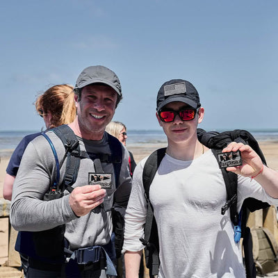 Two people enjoying a day at the beach, both smiling and holding patches. They're each wearing GORUCK backpacks and casual clothing crafted from ToughDry® fabric, under clear blue skies with the ocean behind them. The scene is lively with others partially visible in the background, contributing to the vibrant seaside atmosphere.