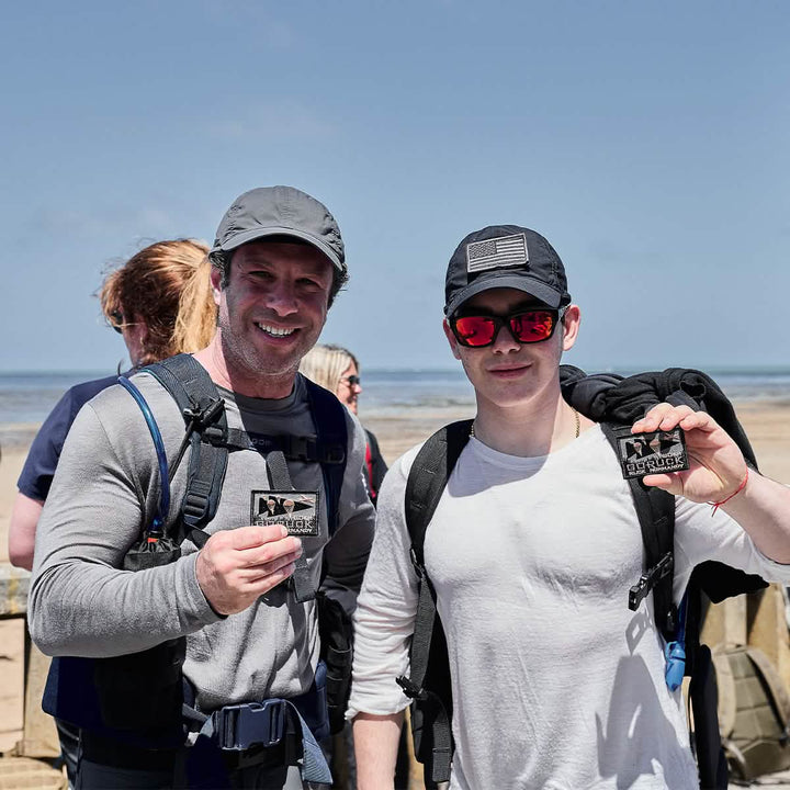 Two people enjoying a day at the beach, both smiling and holding patches. They're each wearing GORUCK backpacks and casual clothing crafted from ToughDry® fabric, under clear blue skies with the ocean behind them. The scene is lively with others partially visible in the background, contributing to the vibrant seaside atmosphere.