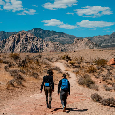 Two adventurers wearing backpacks hike along the desert trail under a blue sky with scattered clouds. Rocky mountains and dry vegetation create a scenic, rugged landscape, while their Trail Cuff Merino Challenge Socks from GORUCK ensure comfort on this wild journey.