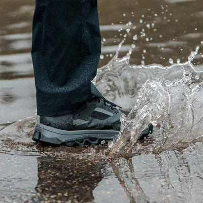Close-up of a person wearing waterproof shoes and dark pants with GORUCK Merino Challenge Socks featuring Trail Cuff detailing, stepping into a puddle as water splashes up. The black shoes boast gray accents and a rugged sole, perfect for outdoor activities.