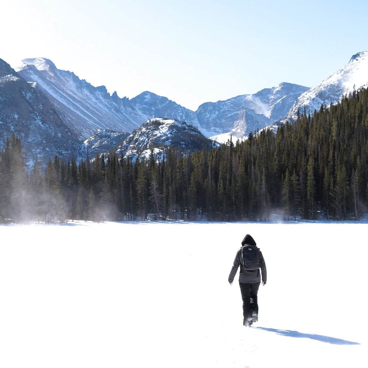 Clad in GORUCK's Merino Challenge Socks - Trail Cuff, a person in winter clothing walks across a snow-covered landscape with snow-capped mountains and a dense forest in the background under a clear blue sky, while mist gently rises from the snow in the bright sunlight.