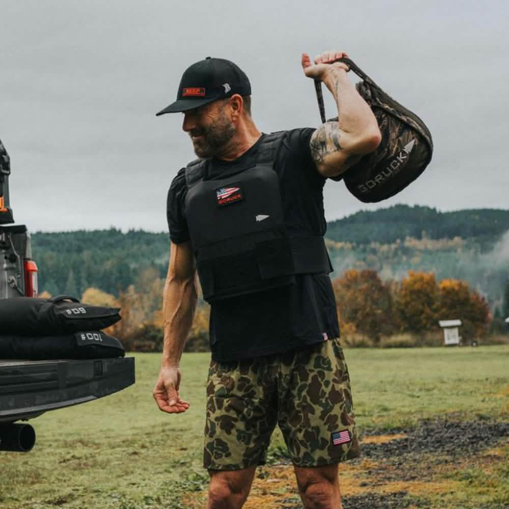 A man in a black cap, black shirt, and camouflage shorts is working out in the open air, sporting the GORUCK Training Weight Vest 2.0. Amidst a grassy field with trees around him, he lifts a sandbag over his shoulder. A truck parked nearby emphasizes his commitment to rigorous training with the adjustable GORUCK vest as part of his routine.