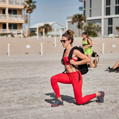 A person in red athletic wear and sunglasses works out on a sandy beach, lunging forward with a backpack. Their GORUCK Women’s Training Leggings Pocket - ToughFlex accentuate the movement. In the background, buildings and palm trees stand under a clear sky.