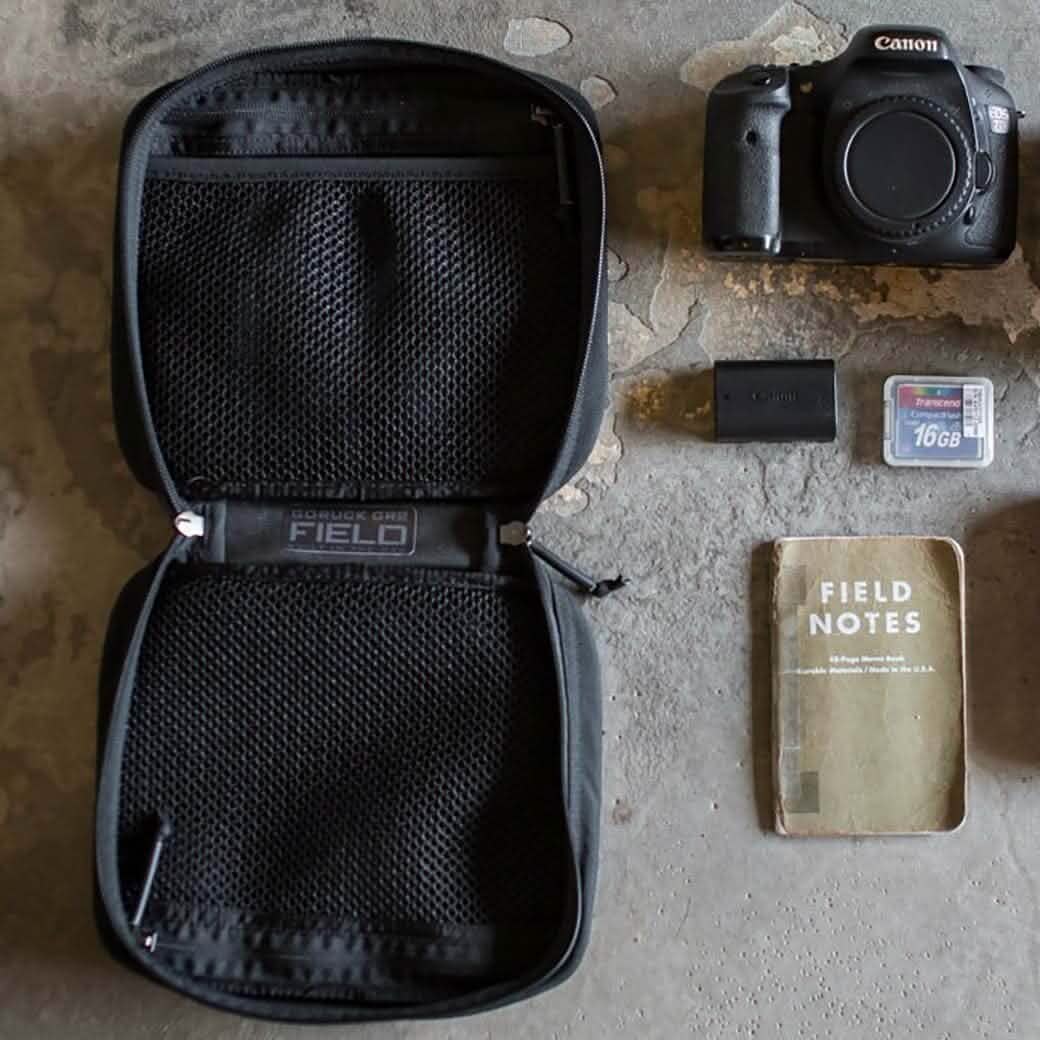 A top-down view of a photography kit on a textured surface includes an open GR2 Field Pocket by GORUCK, featuring MOLLE webbing and mesh compartments. Inside, you'll find a Canon camera body, two memory cards, a camera battery, and a Field Notes notebook neatly arranged like an expert electronics organizer.