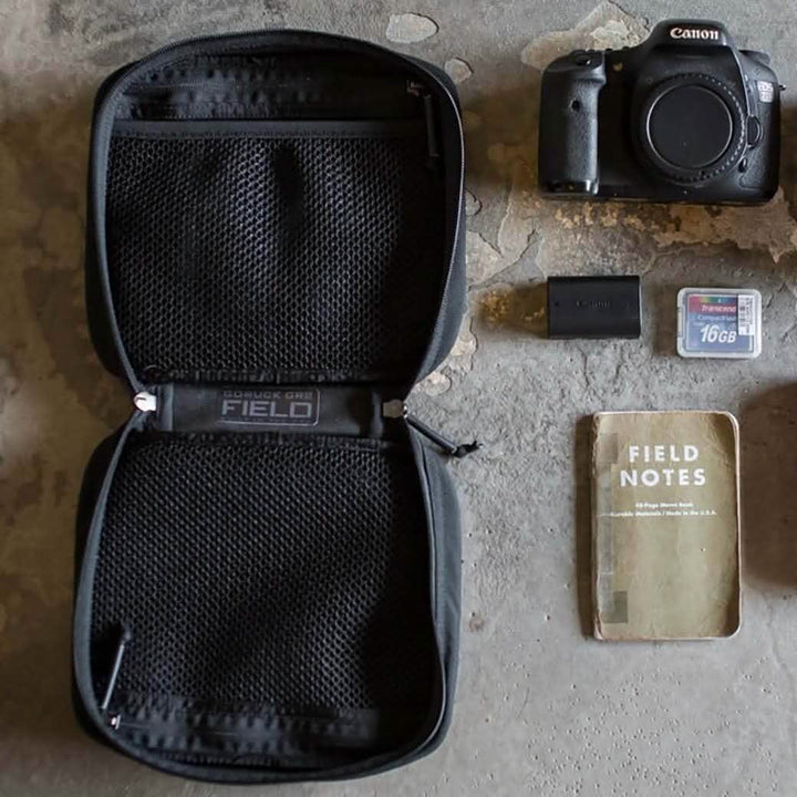 A top-down view of a photography kit on a textured surface includes an open GR2 Field Pocket by GORUCK, featuring MOLLE webbing and mesh compartments. Inside, you'll find a Canon camera body, two memory cards, a camera battery, and a Field Notes notebook neatly arranged like an expert electronics organizer.