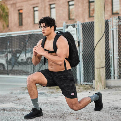Wearing a dark tank top and camo shorts, an individual performs a city street lunge, demonstrating their commitment to functional fitness. With the 3X Support™ from their GORUCK Men's Ballistic Trainers - Blackout w/ Black Reflective Spearhead and a black backpack made of CORDURA® Ballistic Nylon, they stand prepared by the chain-link fence.