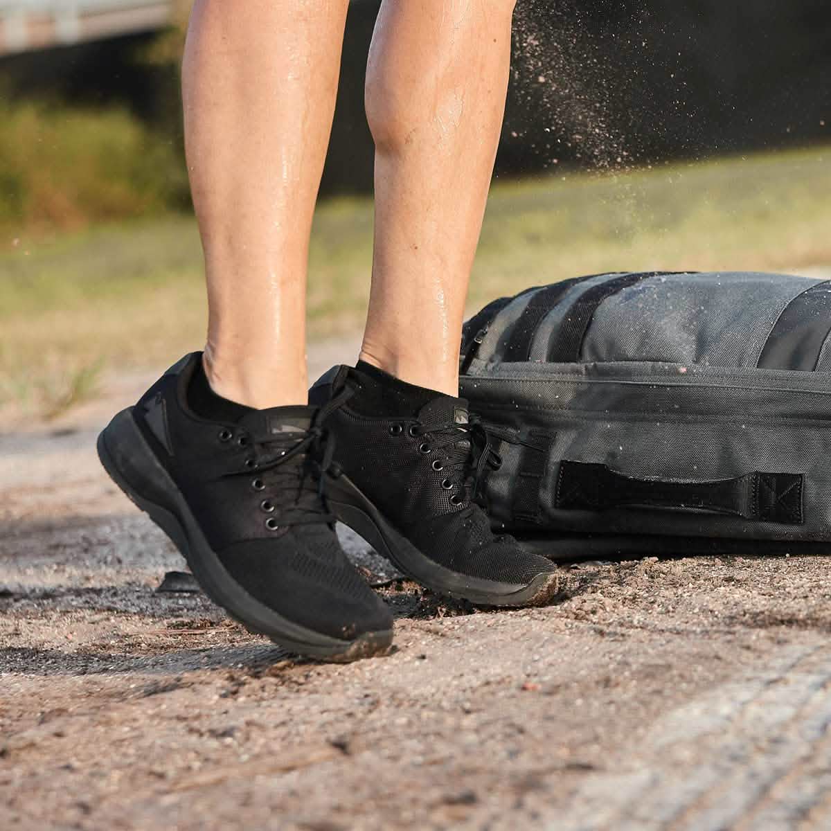 A person wearing GORUCK Men's Ballistic Trainers - Blackout with Black Reflective Spearhead stands near a dusty trail, with a gray backpack on the ground. Dust around their feet suggests movement or jumping, and the blurred background indicates an outdoor setting.