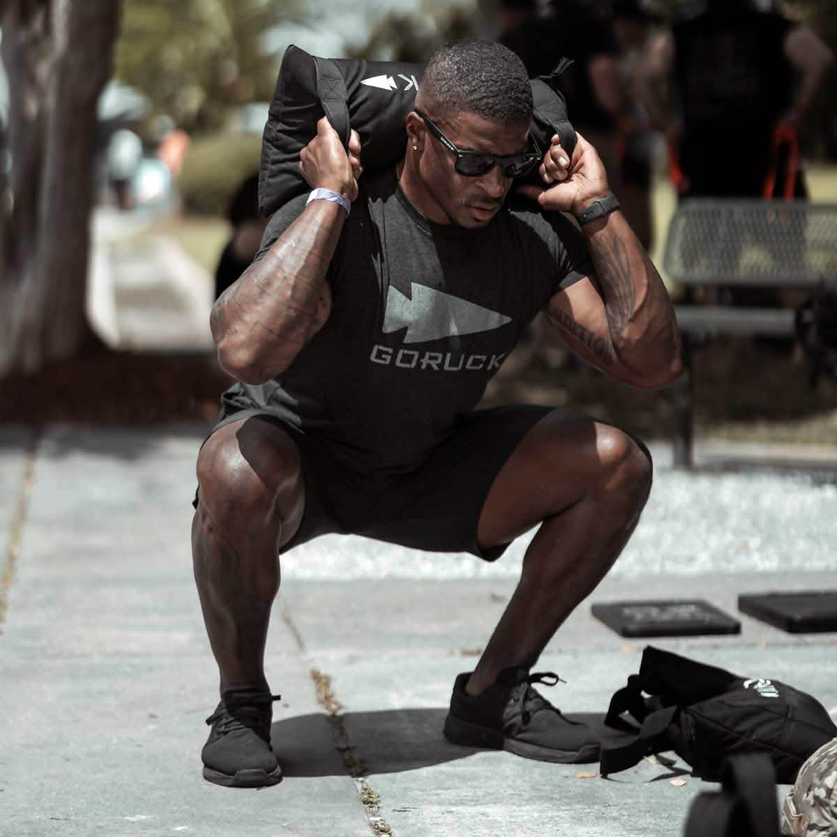 A person wearing sunglasses and a GORUCK t-shirt, showcasing Men's Ballistic Trainers in Blackout with a Black Reflective Spearhead, performs a squat while holding a sandbag on their shoulders. They are outdoors on a paved path, with trees and a bench visible in the background.