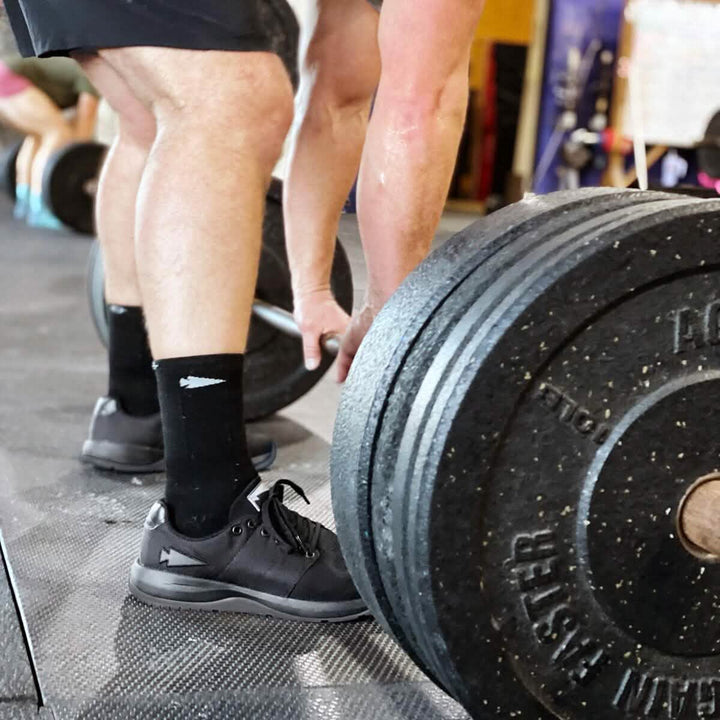 Wearing GORUCK's Men's Ballistic Trainers - Blackout with a Black Reflective Spearhead, a focused athlete prepares to lift a heavy barbell in the gym, highlighting their powerful legs and the weight plates. The scene is set with CORDURA® Ballistic Nylon gear and other dedicated individuals in the background, cultivating an inspiring atmosphere of strength and resilience.