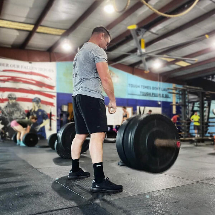 A man in a gym is preparing to lift a heavy barbell, donning GORUCK Men's Ballistic Trainers - Blackout with Black Reflective Spearhead, along with a gray t-shirt and black shorts. Other people can be seen in the background, and a large American flag is painted on the wall.