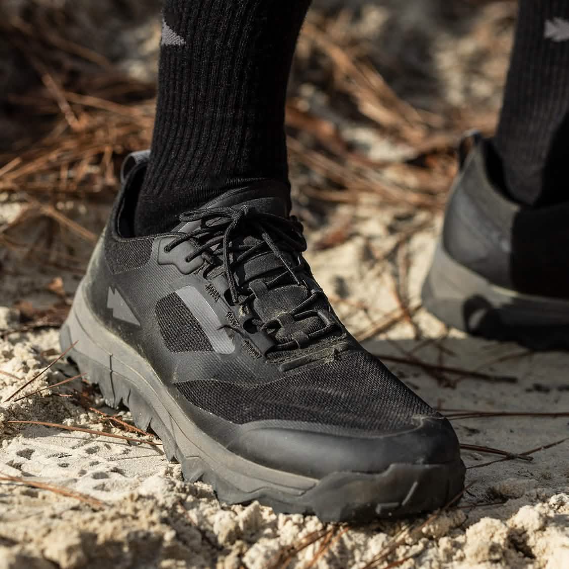 Close-up of a person wearing the Mackall - Black + Forged Iron trail running shoes by GORUCK, ideal for Special Forces training, paired with black socks. They stand on sandy and pine needle-covered ground. The shoes feature a rugged, textured sole, making them perfect rucking shoes for challenging off-road terrains.
